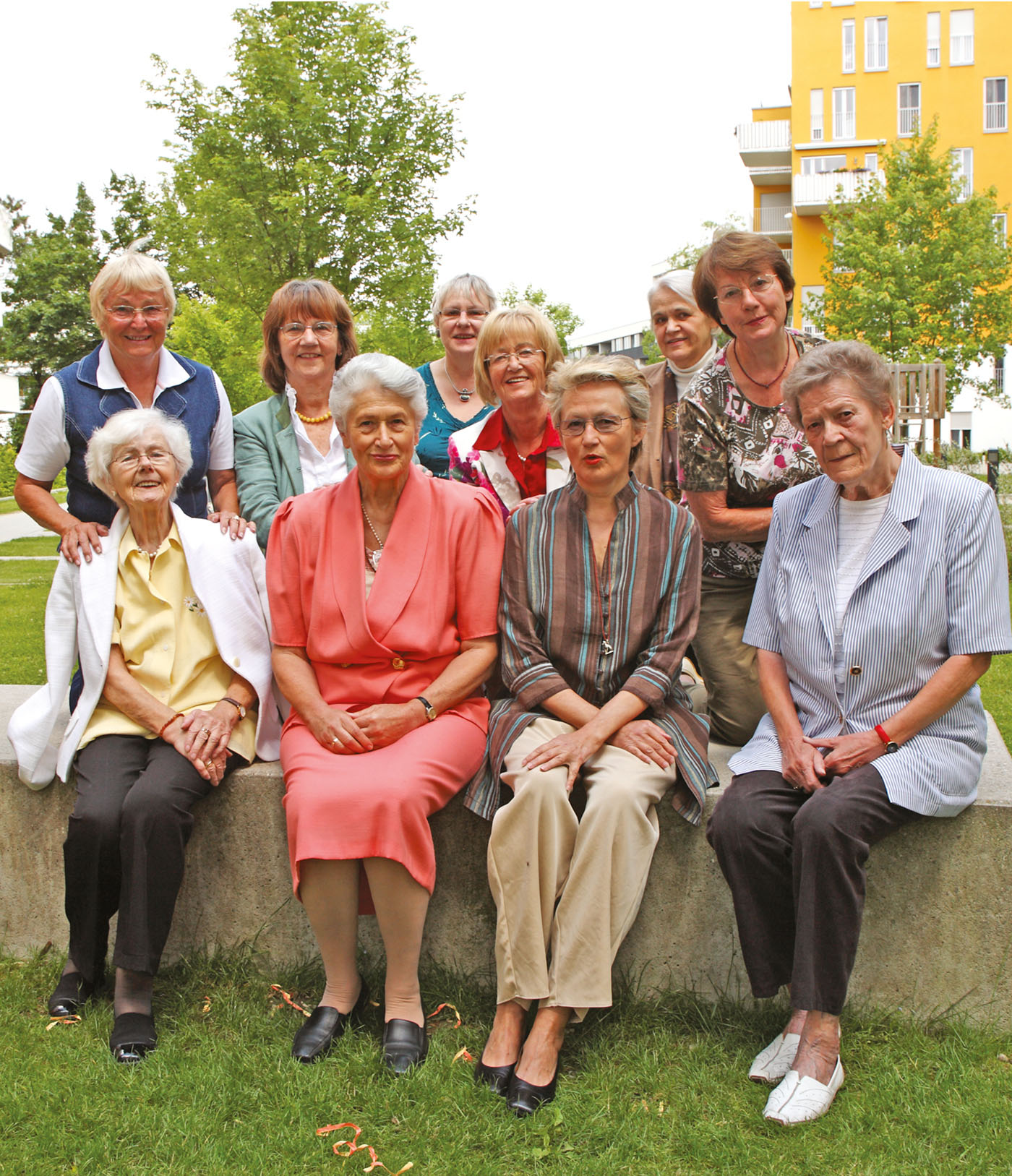 8 Wohnfrauen mit 2 Vorstandsfrauen sitzen  im begrünten Innenhof der Wohnanlage in Vorfreude auf die sorgende Hausgemeinschaft blicken fröhlich in die Kamera